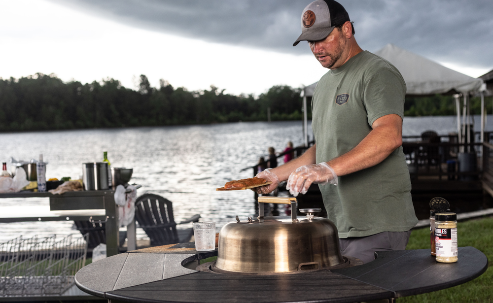 Man grilling food on a rotating charcoal grill by a lake under cloudy sky