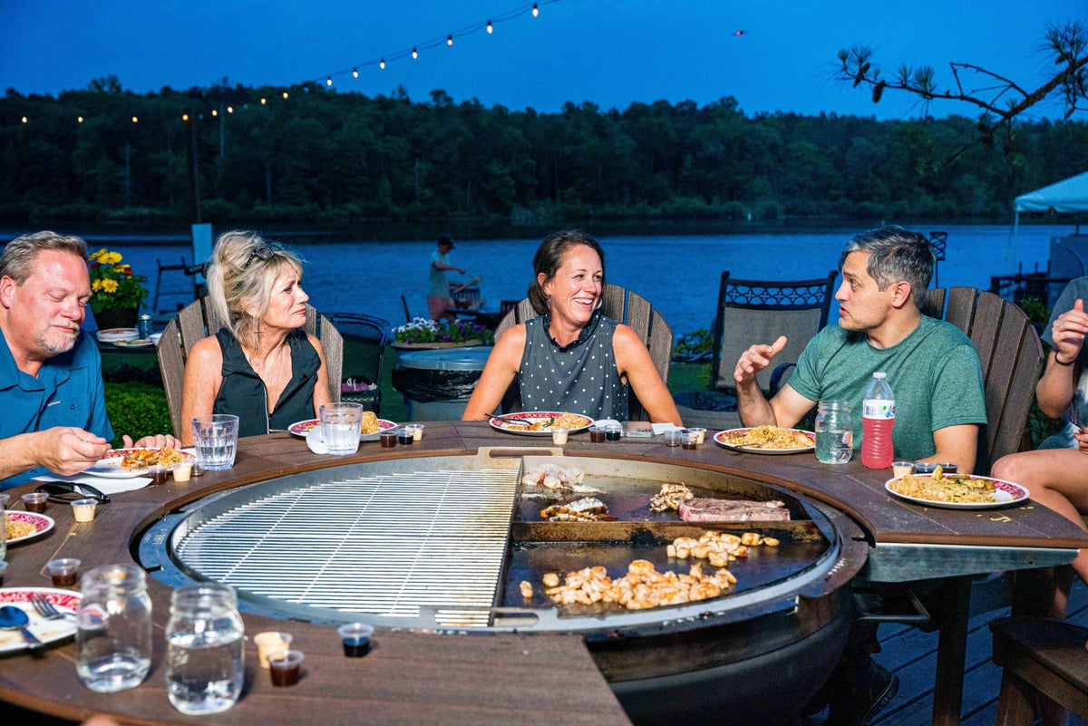 Group enjoying dinner around outdoor 5-in-1 rotating charcoal grill, lakeside in evening.
