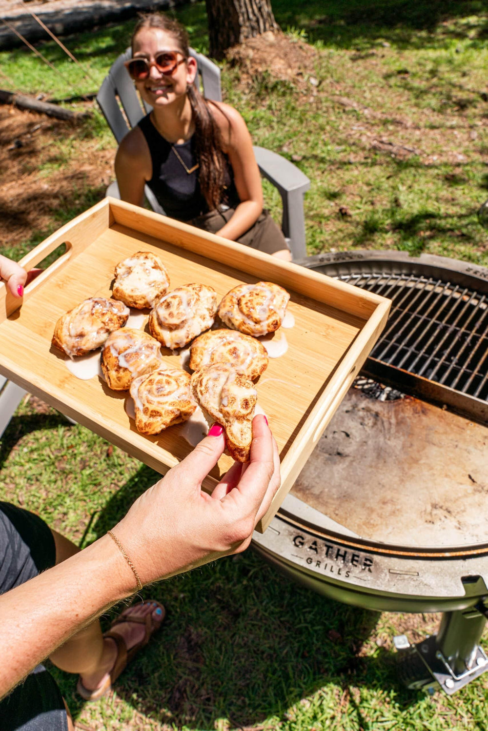 Outdoor charcoal grill with fresh cinnamon rolls on a tray and a woman sitting nearby