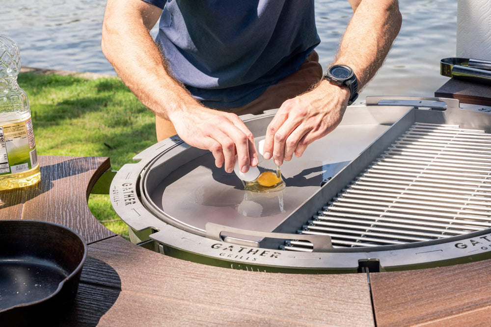 Person cracking an egg onto a stainless steel griddle on a multi-functional outdoor grill by a lake.