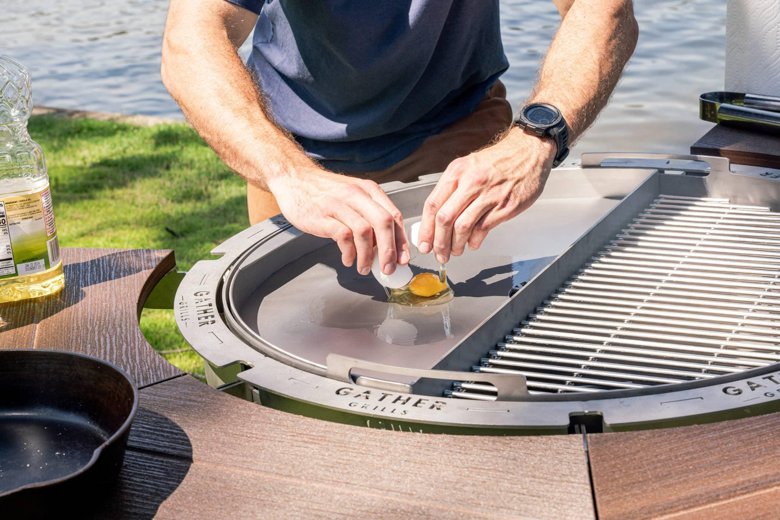 Person cracking an egg onto a stainless steel griddle on a multi-functional outdoor grill by a lake.