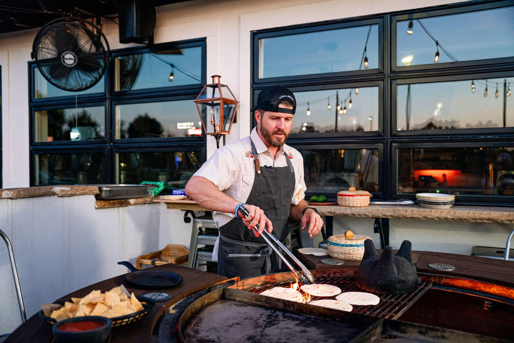 Man grilling tortillas on an outdoor rotating charcoal grill with fire pit at sunset.