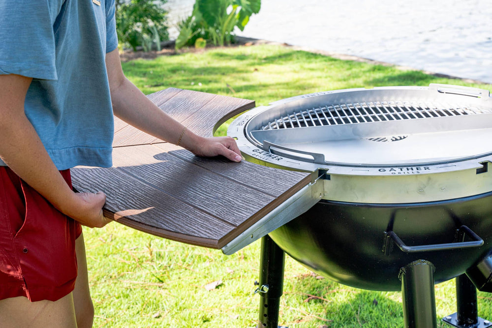 Person assembling round outdoor grill with wooden tabletop by a lake on grass