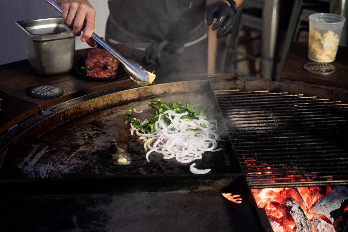 Sliced onions and peppers cooking on a griddle over charcoal grill with glowing coals
