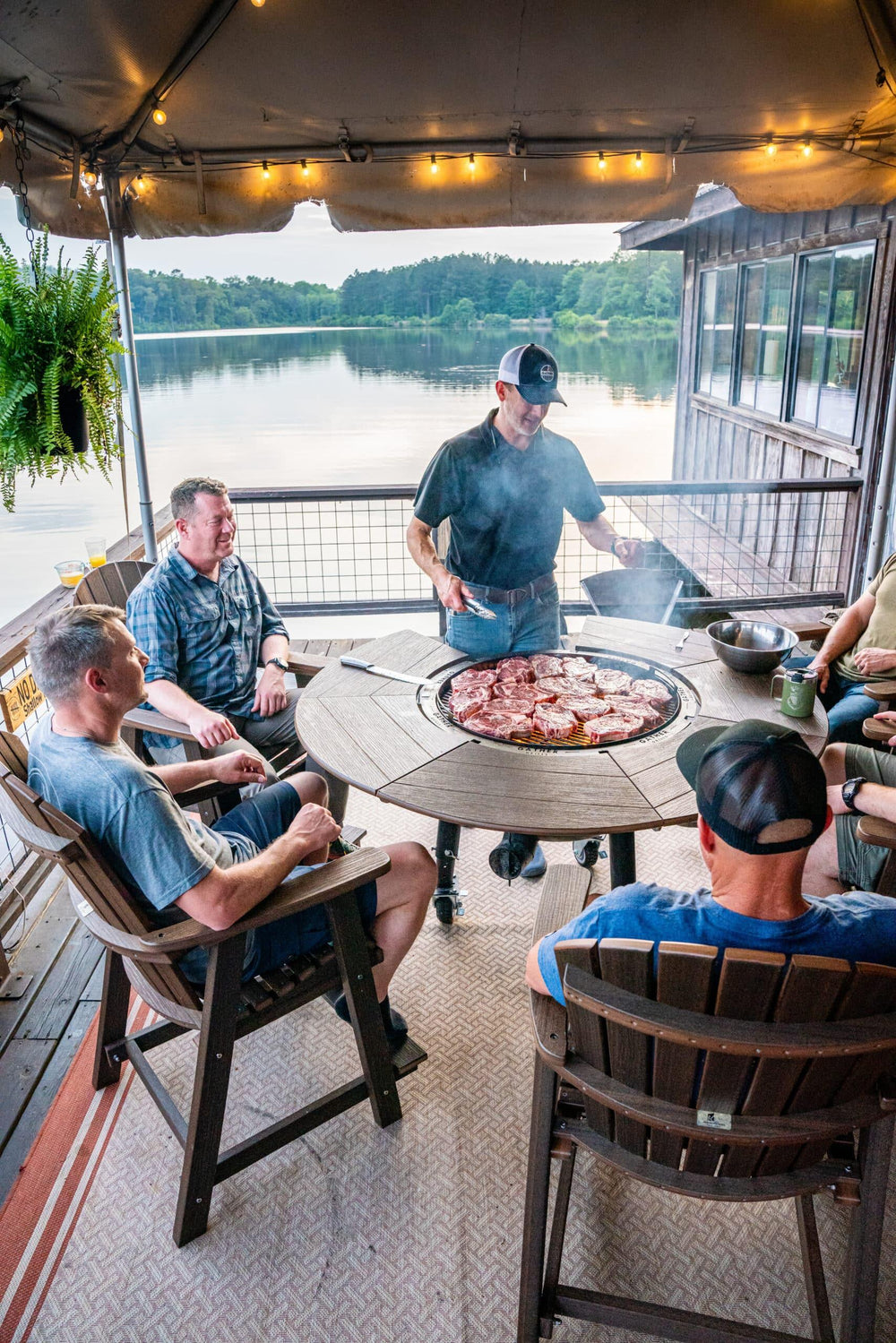 Group of people grilling steaks on a large round outdoor charcoal grill by a lake