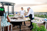 Chefs grilling on a rooftop patio using a large outdoor charcoal grill with dining area at sunset.