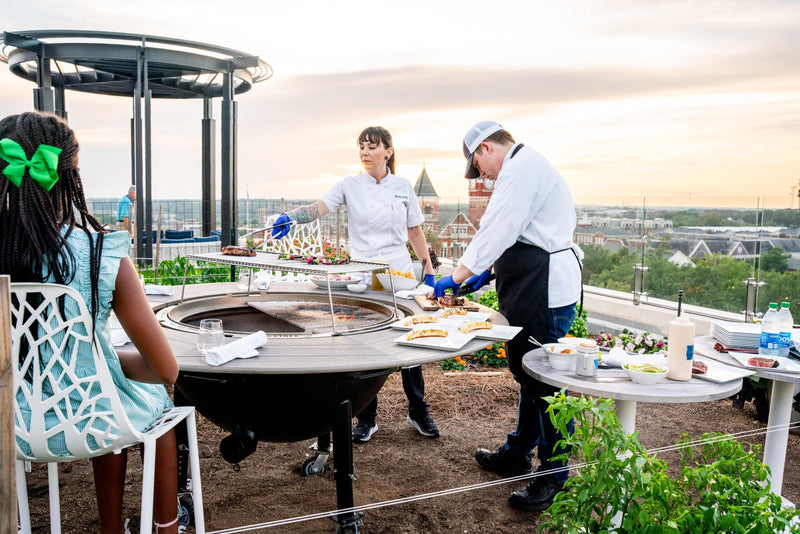 Chefs grilling on a rooftop patio using a large outdoor charcoal grill with dining area at sunset.