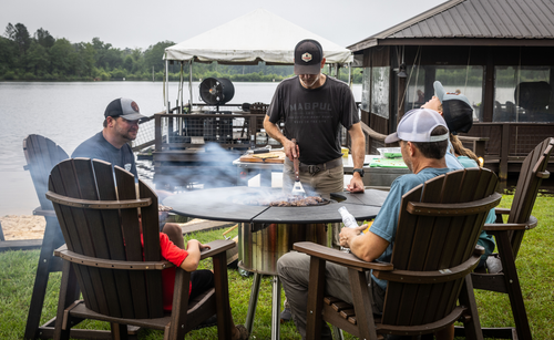 Group enjoying outdoor grilling on a round rotating charcoal grill by a lake