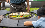 Man grilling corn and vegetables on outdoor rotating charcoal grill surrounded by table seating.