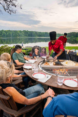 Group enjoying outdoor dining and grilling on a large round charcoal grill by a lake