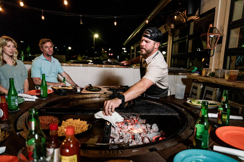 People gathering around a large outdoor charcoal grill and fire pit at night