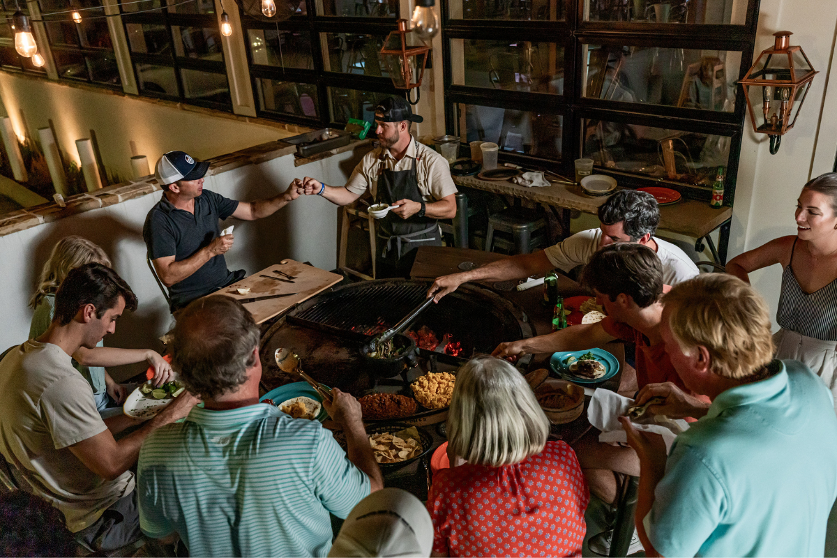 Group gathered around a rotating charcoal grill sharing food and enjoying a social dinner