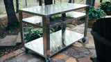 Stainless steel outdoor prep table with wheels on a stone patio, surrounded by plants