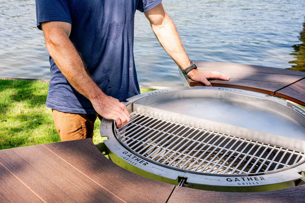 Man adjusting round stainless steel outdoor grill with wood tabletop by a lake