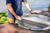 Man adjusting round stainless steel outdoor grill with wood tabletop by a lake