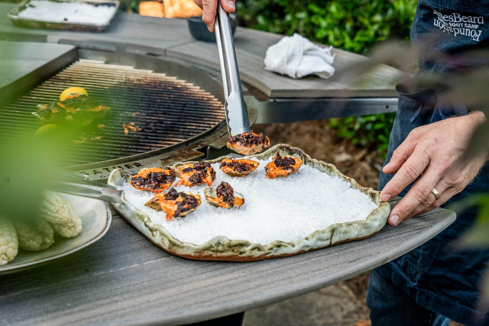 Outdoor grilling oysters on a rotating charcoal grill with tongs and fresh ingredients nearby