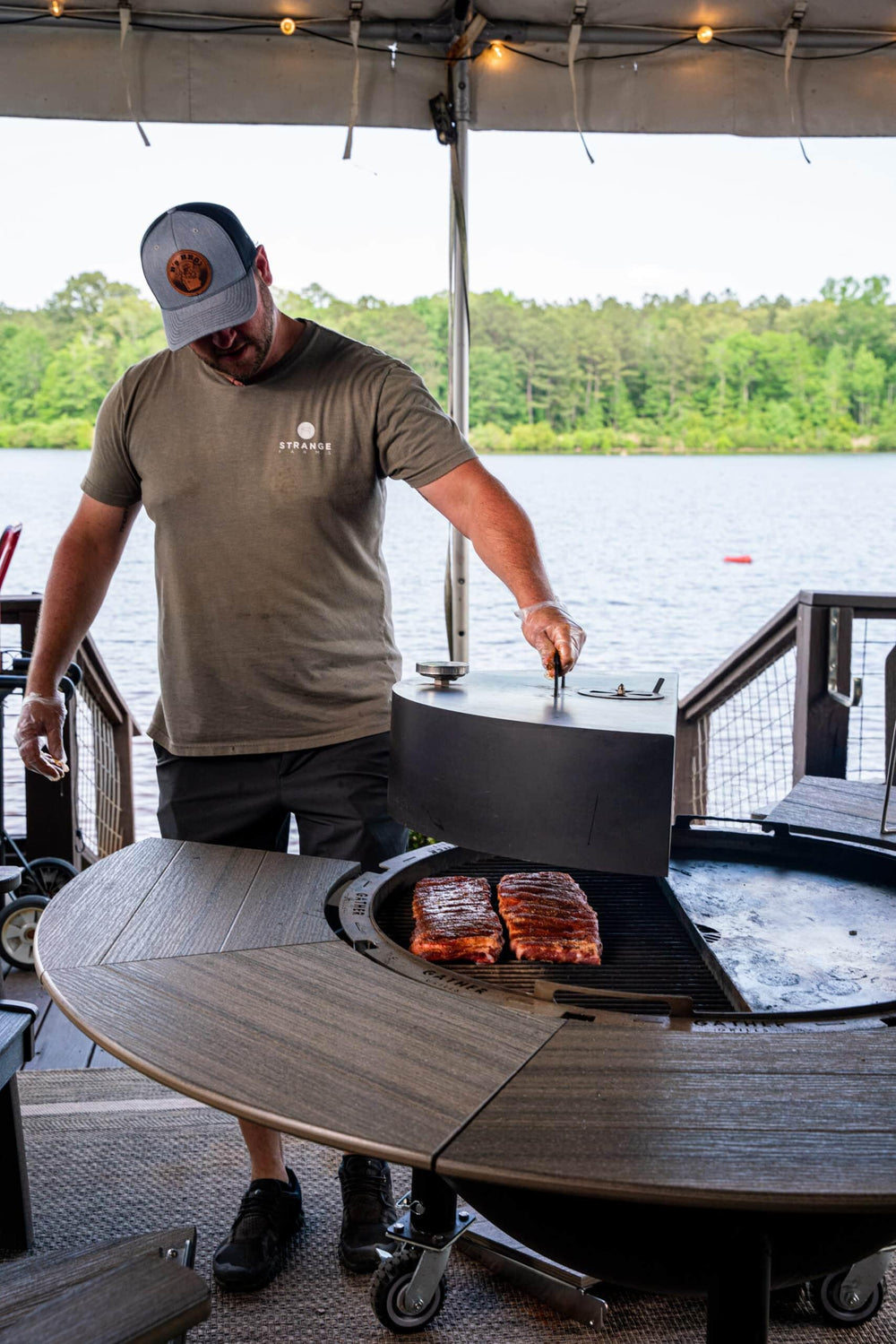 Man grilling ribs on a multi-functional outdoor charcoal grill by a lakeside patio.