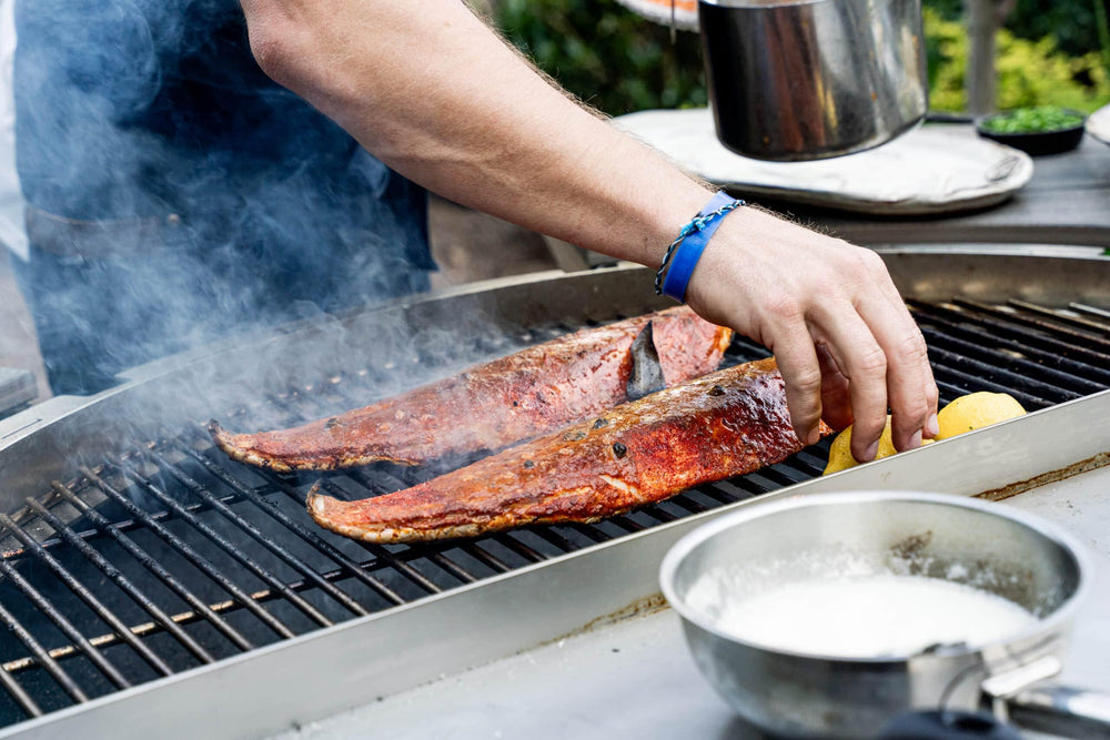 Grilling seasoned fish fillets on a smoky outdoor charcoal grill with a hand adding lemon
